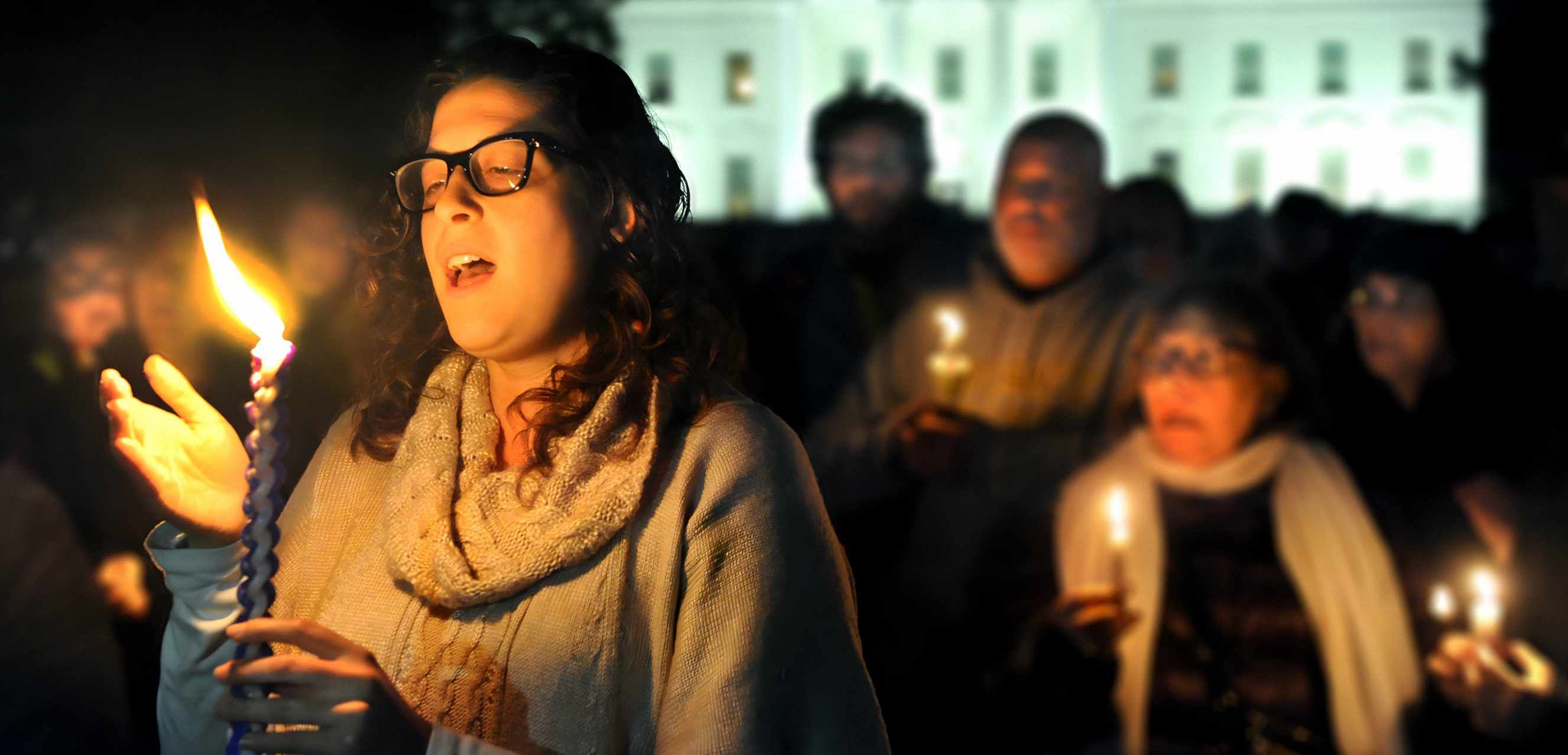 A young woman wearing glasses holds a Havdalah candle with others standing vigilantly in the background, it is night and a government building is lit up in the distance. Her mouth is open as if to sing, her hand is raised to protect the flame.